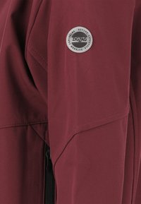 Giacca softshell bordeaux con zip frontale, cuciture oblique e una patch circolare con il logo "ZIGZAG" sulla spalla sinistra.