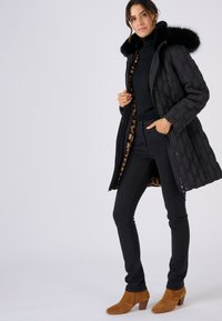 Zwarte gewatteerde jas met een nepbontkraag, voering met luipaardprint, gecombineerd met zwarte jeans en bruine enkellaarsjes met hak, op een witte achtergrond.