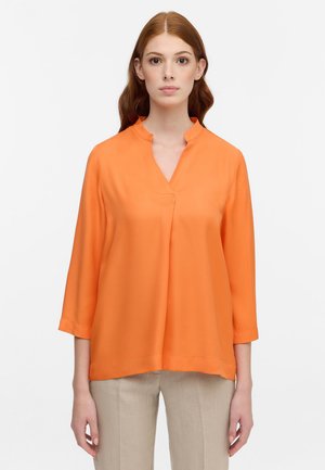 Blusa arancione leggera con scollatura a V e maniche a tre quarti. Texture liscia, vestibilità rilassata e orlo posteriore leggermente più lungo.