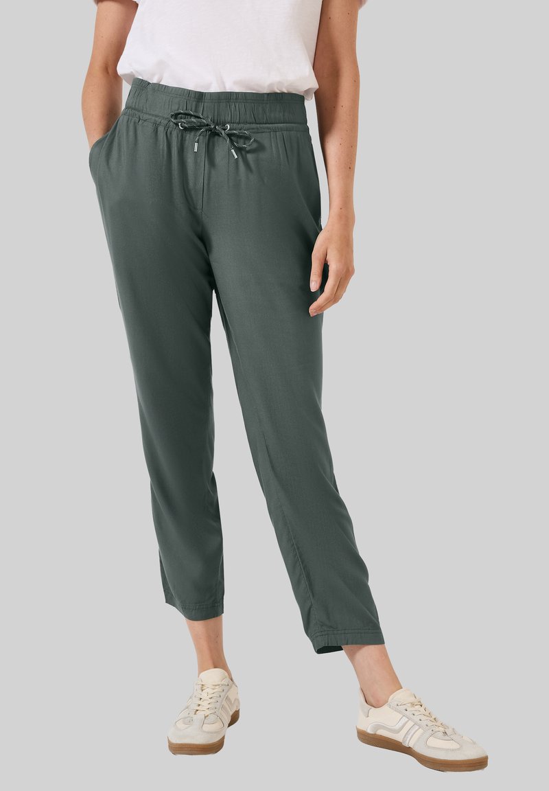 Lightweight, dark green jogger pants feature an elastic waistband with a drawstring, side pockets, and tapered legs, paired with cream sneakers.