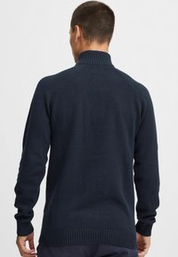 Pull en maille bleu marine avec un col montant, des poignets côtelés et un motif de point texturé, offrant une coupe décontractée et des manches raglan.