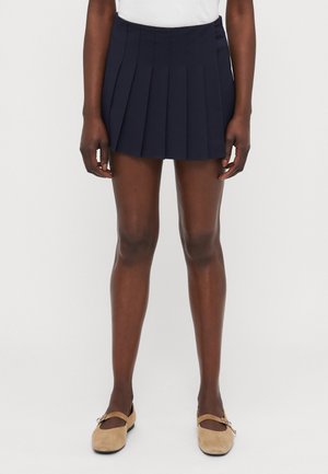 Plisserte marineblå shorts laget av vevd stoff, med høy midje og utsvingt design. Kombinert med lyse beige flate sko.