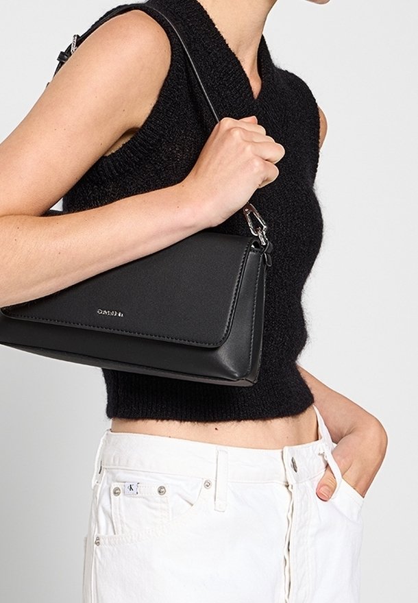 Bolso de mano de cuero negro con diseño estructurado y forma angular. Presenta un logo plateado y una correa desmontable. Llevado en un brazo con pantalones blancos.