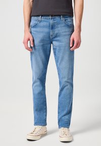 Jeans de mezclilla azul claro con un corte regular, que presentan cinco bolsillos, costuras contrastadas y cierre de botón. Llevados con zapatillas color beige.