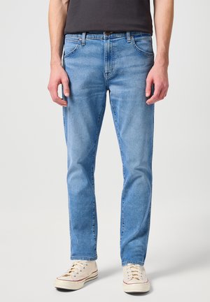 Jeans de mezclilla azul claro con un corte regular, que presentan cinco bolsillos, costuras contrastadas y cierre de botón. Llevados con zapatillas color beige.
