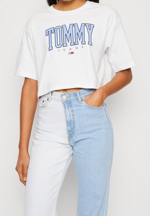 Biała krótka koszulka Tommy Jeans z niebieskim logo w połączeniu z dwukolorowymi jeansami z wysokim stanem, jedna nogawka w jasnym denimu, druga biała.