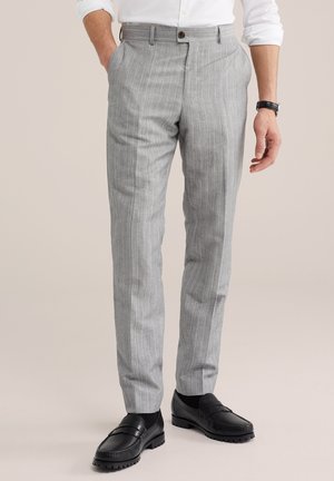 Bărbat purtând pantaloni gri deschis cu dungi fine, pantofi loafer negri și o cămașă albă, stând cu o mână în buzunar pe un fundal neutru.