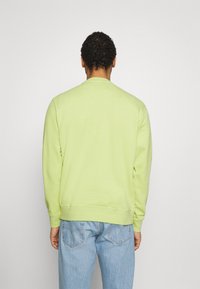 Man i lätt grön långärmad tröja och ljusblå jeans som står med ryggen mot en enfärgad ljusgrå bakgrund.