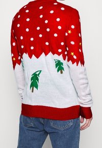 Röd och vit jultröja med gröna träd, vita snöflingor och gula stjärnor, bärs av en person i blå jeans, sett bakifrån.