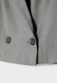 Gros plan d'un blazer en tissu gris clair avec deux boutons noirs et des boutonnières visibles à l'avant.