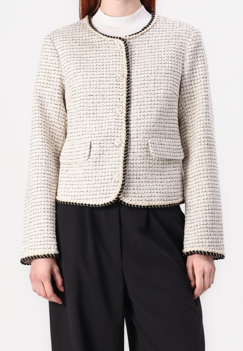 Femme portant une veste en tweed texturée blanche avec des bordures noires, des poches avant et un pantalon noir à jambes larges, debout sur un fond blanc.