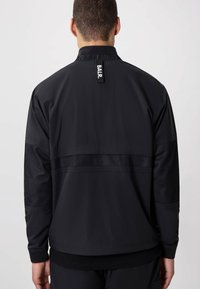 Black jacket with mesh panel accents, featuring a high collar and elastic cuffs. Branding tag visible at the back neckline.