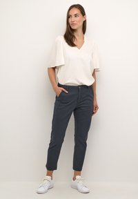 Blouse crème clair avec des manches flutter, associée à un pantalon bleu foncé bien ajusté. Des baskets blanches viennent compléter la tenue.