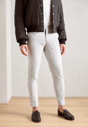 Hellgraue, maßgeschneiderte Hose mit feiner Textur, schmaler Passform und seitlichen Schlitzen, kombiniert mit dunkelbraunen Slippern und einer schwarzen Bomberjacke.