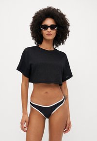 Maglietta nera corta con maniche corte, realizzata in un tessuto testurizzato. Abbinata a un bikini bottom nero rifinito in bianco, creando un look a contrasto.