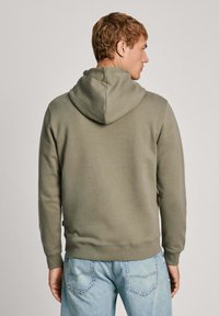 Sudadera verde con capucha, de textura suave, con puños y dobladillo acanalados. Se lleva con jeans de un azul claro, mostrando un ajuste relajado desde la parte trasera.