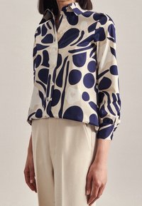 Collared blouse in cream and navy, featuring organic shapes and polka dots. Long sleeves with gathered cuffs; fabric has a smooth texture.