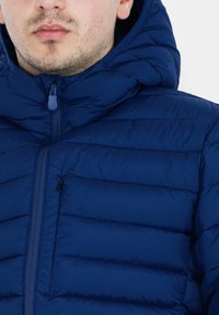 Giacca puffer blu navy con sezioni imbottite, chiusura zip e cappuccio. Presenta una piccola tasca con zip sul lato sinistro del petto. Texture liscia.