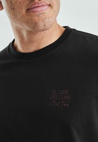 Czarny bawełniany T-shirt z okrągłym dekoltem. Na przodzie widnieje napis w kolorze bordowym: "WYGLĄDAM JAKOŚ ZAINTERESOWANY Dobry Czas." Gładka tekstura.