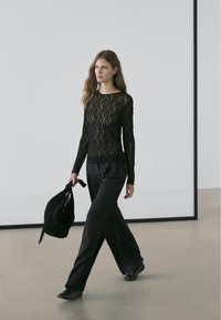 Black lace long-sleeve top with a textured pattern, paired with loose black trousers, and carrying a black bag. Flat shoes complete the look.