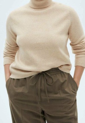 Personne portant un pull à col roulé beige rentré dans un pantalon vert olive à cordon avec les mains dans les poches.