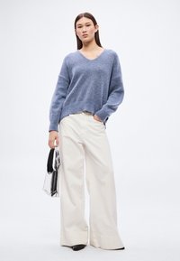 Ανοιχτό μπλε πλεκτό πουλόβερ με V-λαιμόκοψη, συνδυασμένο με εκρού παντελόνια με φαρδύ πόδι. Το μοντέλο κρατά διαφανή τσάντα. Ομαλή υφή συνολικά.