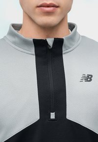 Κοντινό πλάνο ενός ατόμου που φοράει γκρι και μαύρο αθλητικό μπλουζάκι με φερμουάρ και ψηλό γιακά, με το λογότυπο της New Balance στο στήθος.
