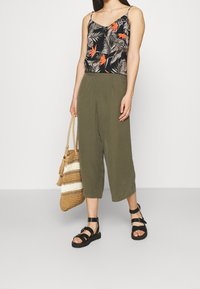 Black floral tank top with orange accents, olive green wide-leg trousers, black strappy sandals, and woven straw bag with striped detail.
