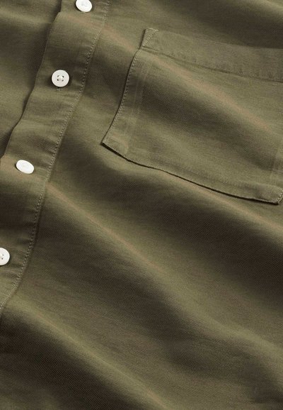 Oliven grøn stofskjorte med en blød tekstur, udstyret med hvide knapper og en brystlomme. Diskret syning giver visuel interesse.