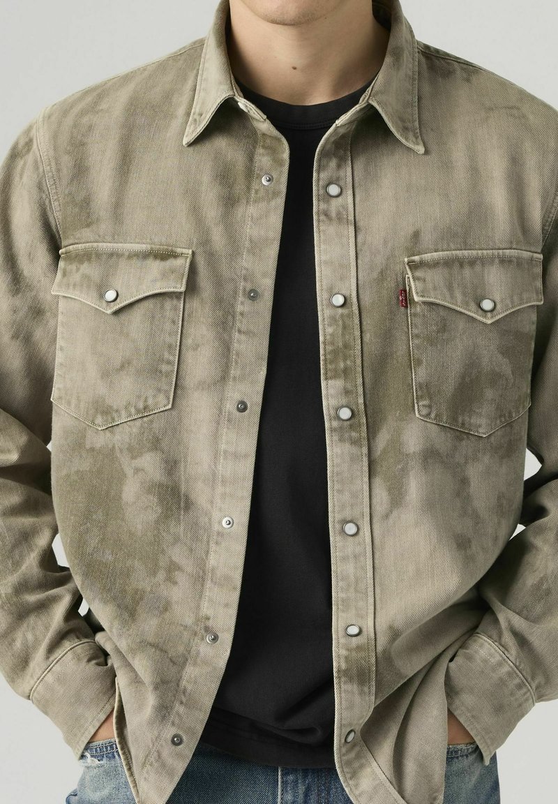 Man wearing a faded olive green button-up shirt over a black t-shirt with hands in jean pockets, cropped at shoulders and waist.