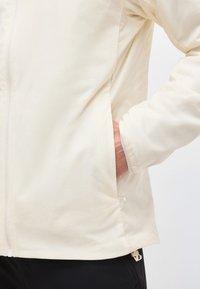 Personne portant une veste écrue avec une main dans la poche latérale, montrant une partie de la manche et un pantalon noir.