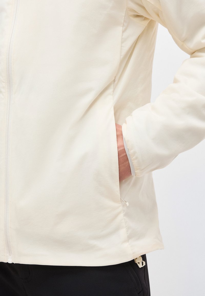 Persona che indossa una giacca color avorio con una mano nella tasca laterale, che mostra parte della manica e pantaloni neri.