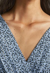 Vaaleansininen mekko, jossa on V-pääntie, musta-valkoisella kukkakuviolla ja hienovaraisilla kultaisilla yksityiskohdilla. Sujuva voitteinen pinta, joka kietoutuu vartalon ympäri.