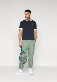 Μπλε ναυτικό πόλο πουκάμισο με λευκή λεπτομέρεια και λογότυπο, συνδυασμένο με ανοιχτό πράσινο αθλητικό παντελόνι. Κρατά ρακέτα τένις και φοράει λευκά αθλητικά παπούτσια.