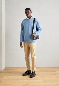 Man in een lichtblauwe sweatshirt, beige broek, zwarte schoenen en een zwarte schoudertas staat op een houten vloer tegen een plain muur.