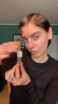 Montre-bracelet en métal doré avec un cadran rectangulaire, un cadran argenté et des accents semblables à des diamants. Attachée à un bracelet à maillons doré.