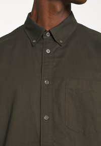 Camicia abituale verde scuro con colletto classico, tasca a protezione e dettagli con bottoni nascosti. Realizzata in tessuto leggero con una superficie liscia.