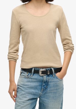 Person wearing a beige long-sleeve top and light blue jeans with a black belt with a decorative silver buckle, one hand in pocket.