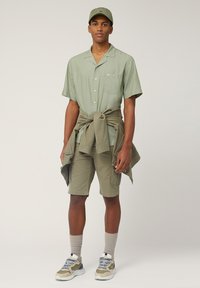 Camisa de manga corta de color verde claro con un bolsillo, combinada con pantalones cortos a juego y una chaqueta atada. Completada con calcetines gris claro y zapatillas.