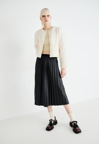 Krämfärgad fleecejacka med knappar, en kort beige topp, plisserad svart midikjol och blanka svarta skor med rem och grov sula.