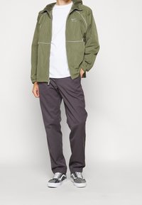 Olivgrön lättviktsjacka med vita detaljer, i kombination med grå byxor och grå mönstrade sneakers. Enkelt design och avslappnad passform.