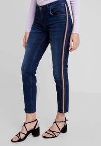 Mörkblå denimjeans med slim fit, med ett bronsrand längs den yttre sömmen och fransade nederkanter vid anklarna. Bärs med svarta remsandaler.