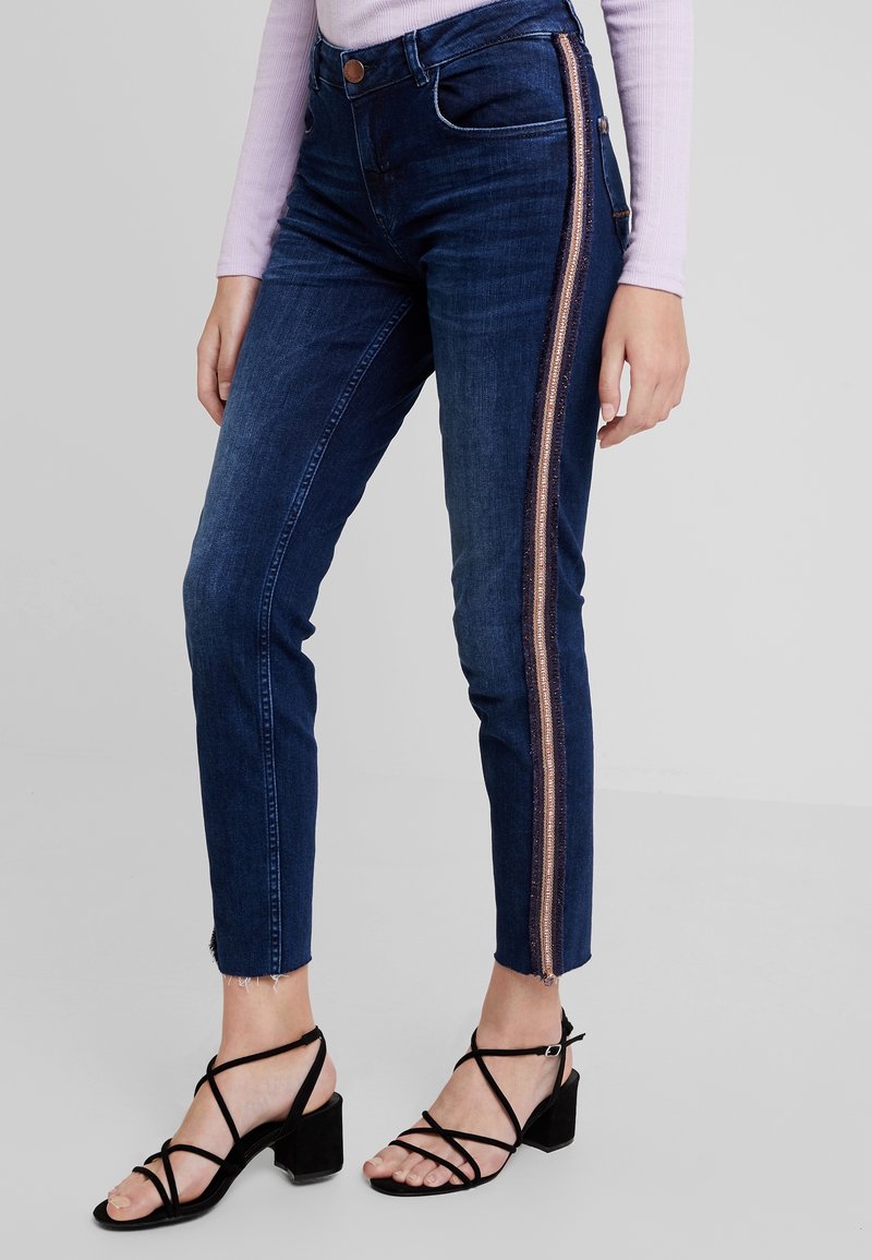 Mörkblå denimjeans med slim fit, med ett bronsrand längs den yttre sömmen och fransade nederkanter vid anklarna. Bärs med svarta remsandaler.