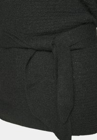 Top negro de punto con un tejido texturizado, detalle de cintura atada y corte holgado. El material parece suave y ligeramente elástico.