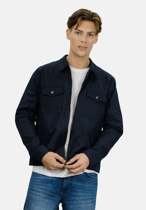 Ung mann iført en marineblå jakke over en hvit t-skjorte og blå jeans, som fester glidelåsen på jakken, mot en ensfarget hvit bakgrunn.