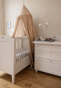 Patut alb din lemn cu laterale din barete, baldachin bej și lenjerie de pat gri. Lângă acesta, o comodă albă cu accente aurii și un carusel decorativ.