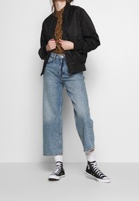 Černá bomber bunda, hnědý vzorovaný top, světle modré široké džíny a černé tenisky s vysokým střihem a bílými podrážkami.