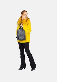 Cappotto giallo con colletto in pelliccia sintetica, pantaloni neri svasati e stivali con tacco. Tiene uno zaino nero a motivo con chiusura a coulisse.