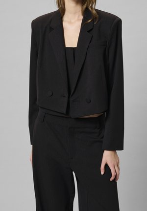 Kvinde iført en sort cropped blazer med matchende bukser med højt talje mod en ensfarvet grå baggrund.