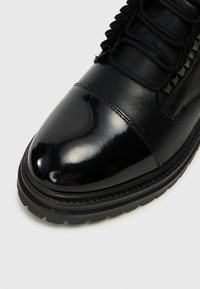 Bota de cuero negra con una puntera brillante, cordones texturizados y una suela resistente. Material flexible con un detallado acabo de costura.